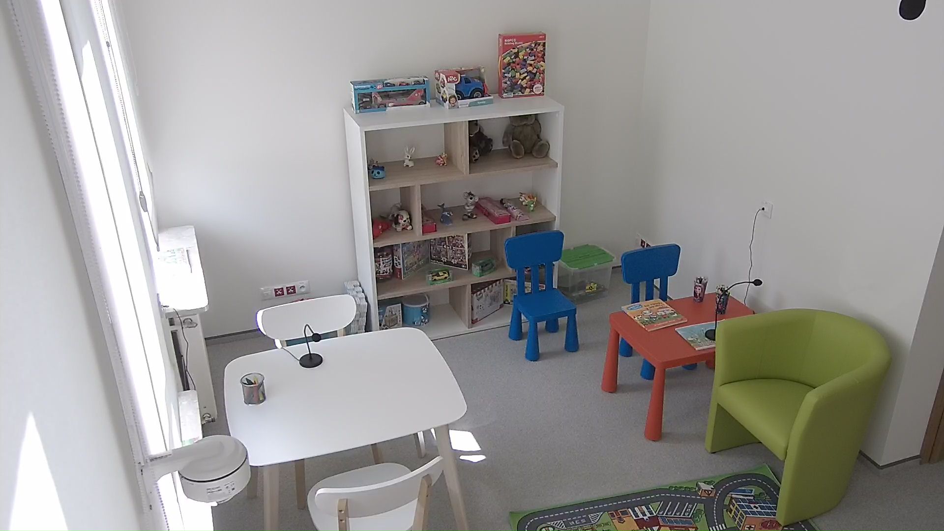 Zdjęcie przedstawia przyjazny pokój przesłuchań. Po lewej stronie biały stolik z 2 białymi krzesłami stojącymi naprzeciwko siebie, po dwóch stronach stolika. Stolik i krzesła są w rozmiarze umożliwiającym siedzenie osobom dorosłym. Po prawej stronie czerwony stolik dla małych dzieci i 2 niebieskie krzesełka dostosowane rozmiarem do stolika. Przed stolikiem stoi zielony fotel w rozmiarze umożliwiającym siedzenie osobom dorosłym. Między stolikami znajduje się przestrzeń umożliwiająca przejście. Z tyłu przy ścianie znajduje się regał z 4 półkami na których ustawione są różne zabawki.